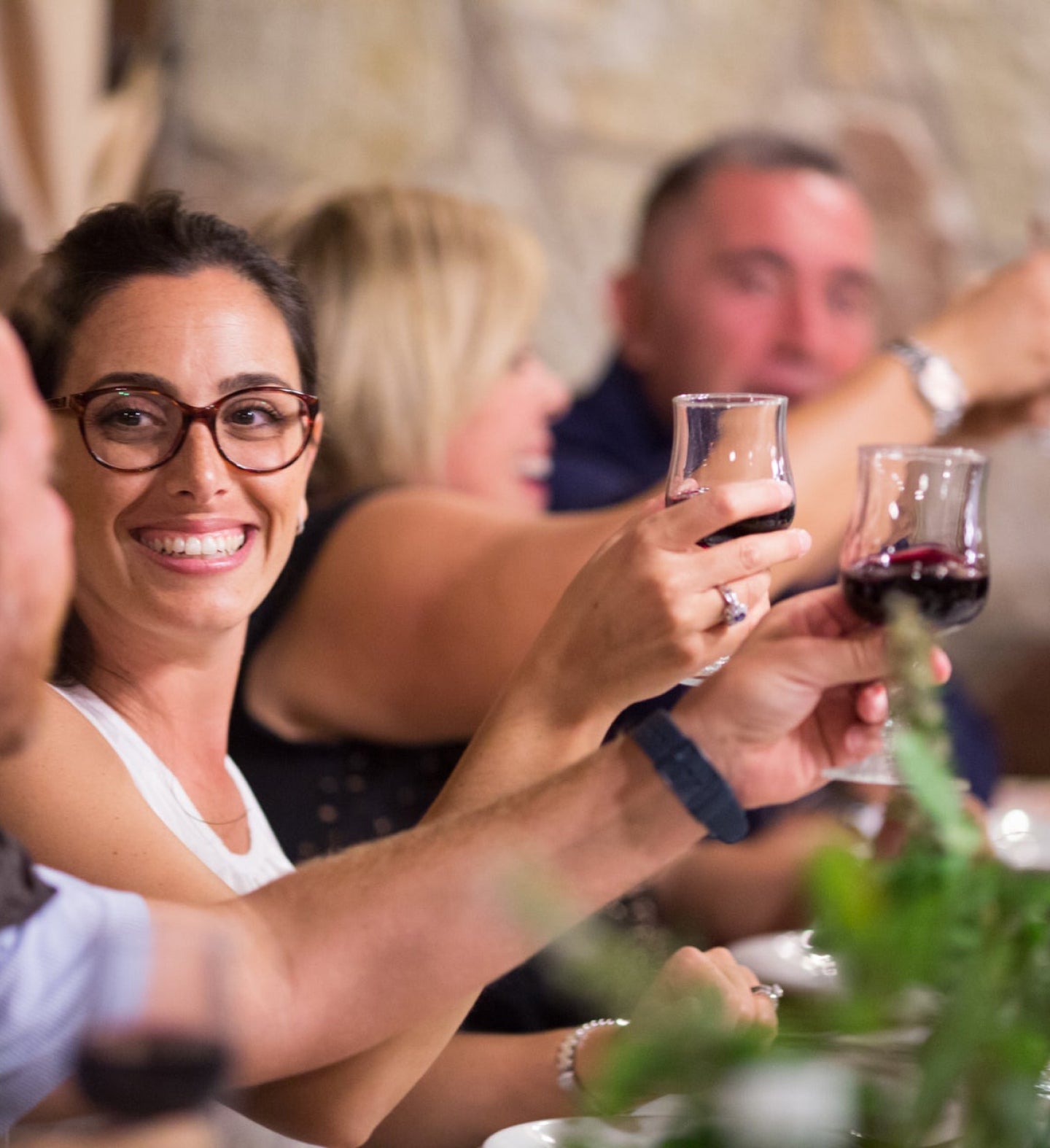 A woman smiling and raising a glass of red wine in a celebratory toast, with other people visible in the background, enjoying the moment